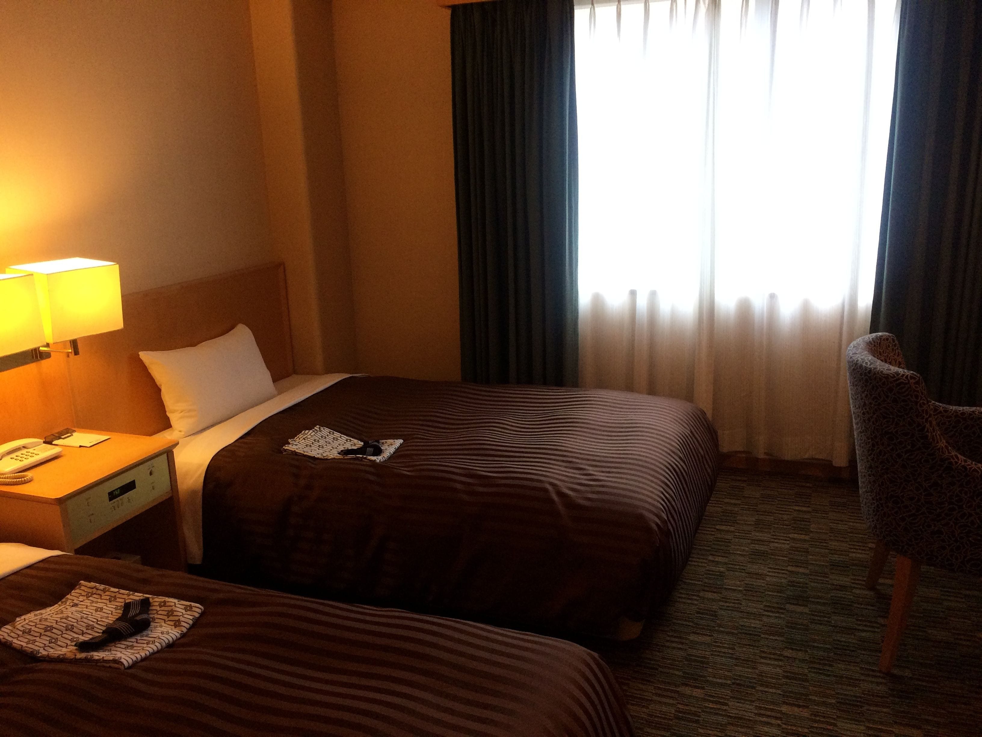 Habitació d'hotel amb dos llits, una tauleta de nit amb una làmpada, una cadira i una finestra amb cortines.