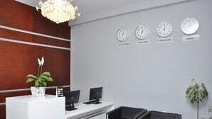 A reception area with a white desk, two black chairs, a potted plant, and clocks displaying times for Addis Ababa, New York, Hong Kong, and Paris. - Afroaddis Hotel Apartment (Addis Ababa)