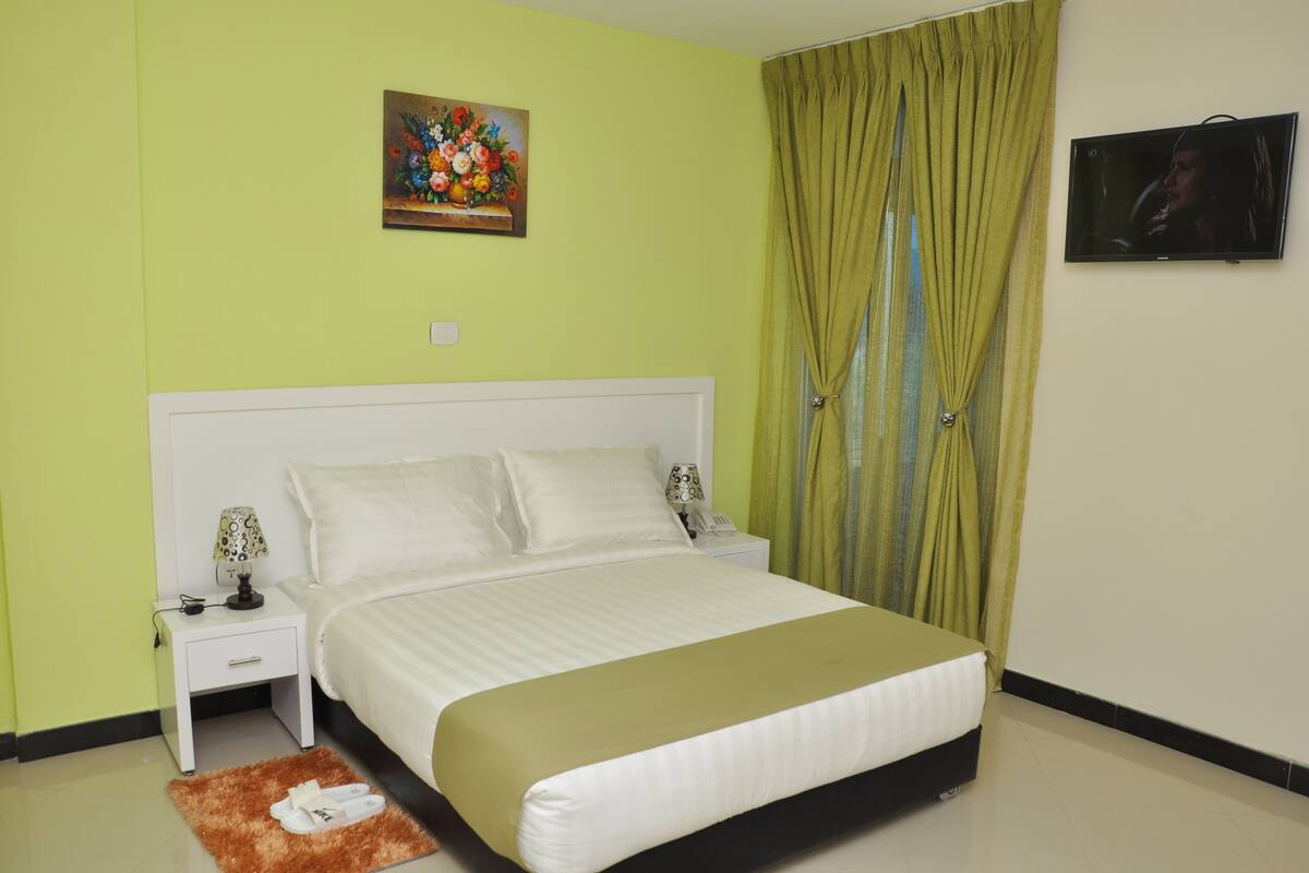 A bedroom with a bed, nightstand, lamp, slippers, and a painting on the wall.