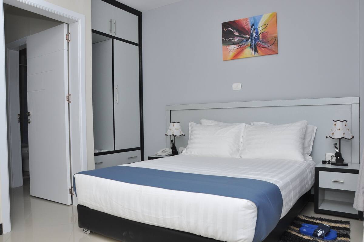 A hotel room with a bed, bedside tables, a wardrobe, and a painting on the wall.