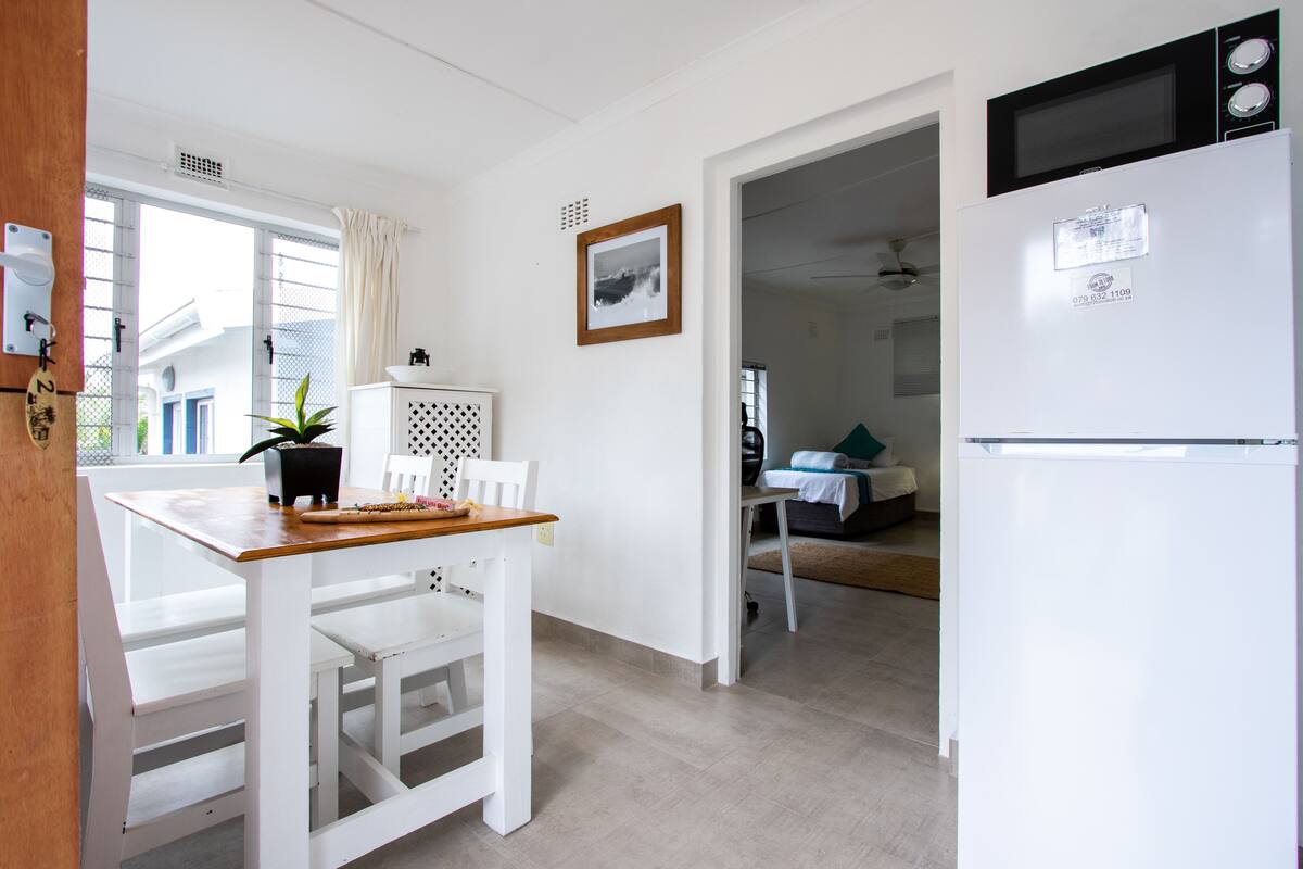 A small, bright kitchen with a white refrigerator, a microwave, and a dining table with chairs.