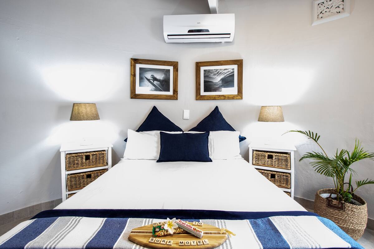 A neatly arranged bed with a blue and white striped bedspread, two framed pictures on the wall, and a welcome mat with a palm tree and a bottle of water.
