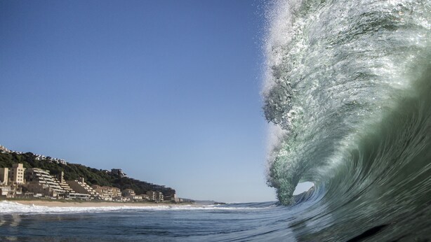 A coastal view with a large wave curling over the water, buildings on the shore, and a clear sky. - Surf Motel (eMdloti)