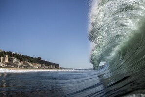 A coastal view with a large wave curling over the water, buildings on the shore, and a clear sky. - Surf Motel (eMdloti)