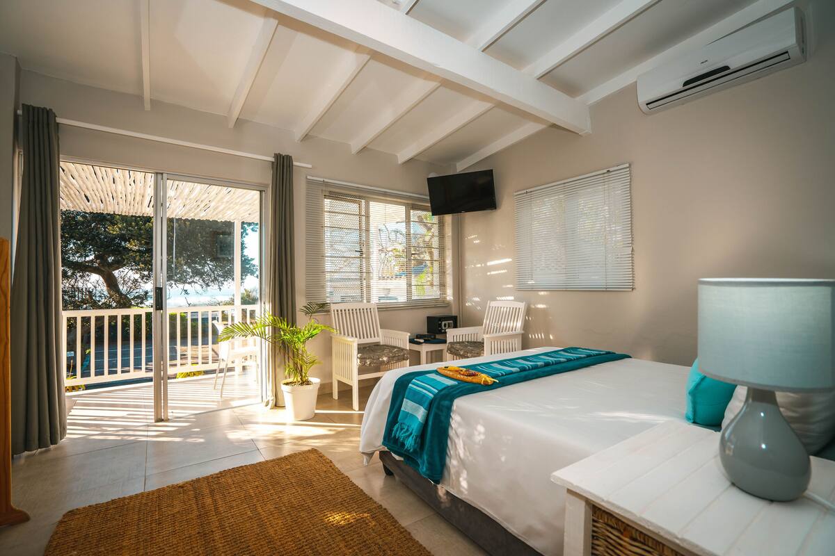 A bedroom with a bed, a desk, a chair, a TV, and a balcony with a potted plant.