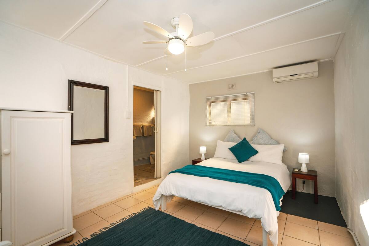 A bedroom with a bed, a ceiling fan, a window with blinds, a bedside table with a lamp, and a door leading to a bathroom.