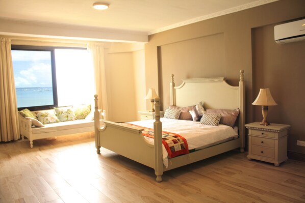 A bedroom with a four-poster bed, a nightstand with a lamp, a bedside table, and a view of the sea through a large window.