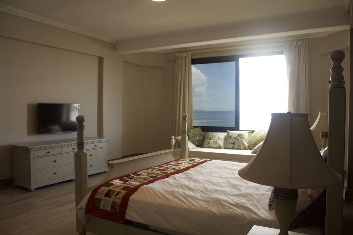 Een slaapkamer met een bed, een dressoir, een televisie en een raam met uitzicht op zee.