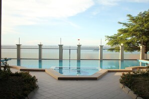 A swimming pool with a tiled deck, surrounded by a metal fence, and a view of the sea in the background.