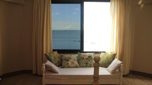 Ein Zimmer mit Meerblick durch ein großes Fenster, ein weißes Sofa mit floralen Kissen und ein hölzernes Kopfstück.