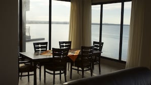 Ein Essbereich mit Tisch und Stühlen, große Fenster mit Vorhängen und Blick auf ein Gewässer.