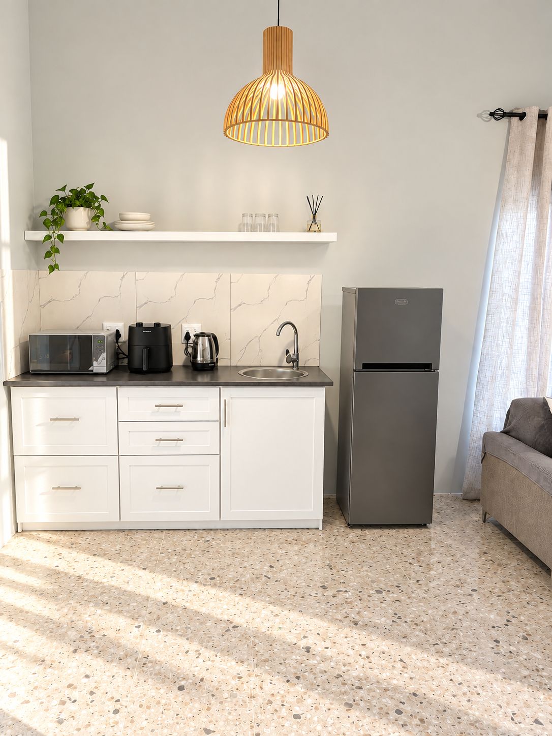 Une cuisine moderne équipée d’un four à micro-ondes encastré, d’une bouilloire et d’un réfrigérateur, d’un luminaire suspendu et d’une plante posée sur le plan de travail.