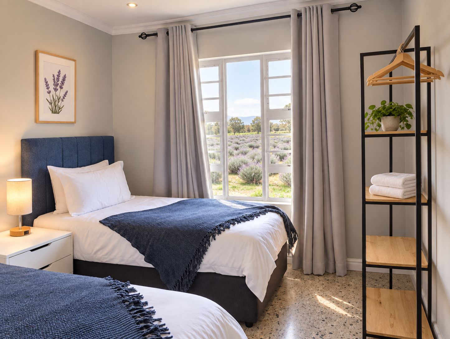 A hotel room with a bed, bedside table, lamp, window with curtains, and a shelf with towels and a plant.