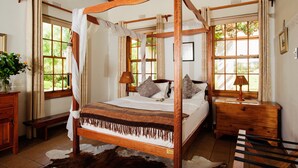 Una cama con dosel, mesitas de noche de madera, un banco y un ventanal con cortinas.