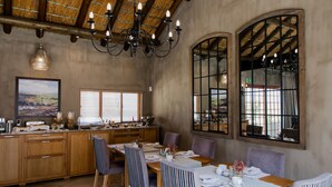 Un comedor amplio con una larga mesa de madera, varias sillas y una araña. Se observa una zona de cocina con armarios de madera y una ventana con persianas.