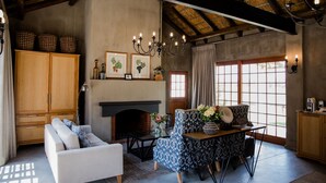 Una sala de estar acogedora con chimenea, un sofá, un sillón estampado y una mesa de comedor con un jarrón de flores.