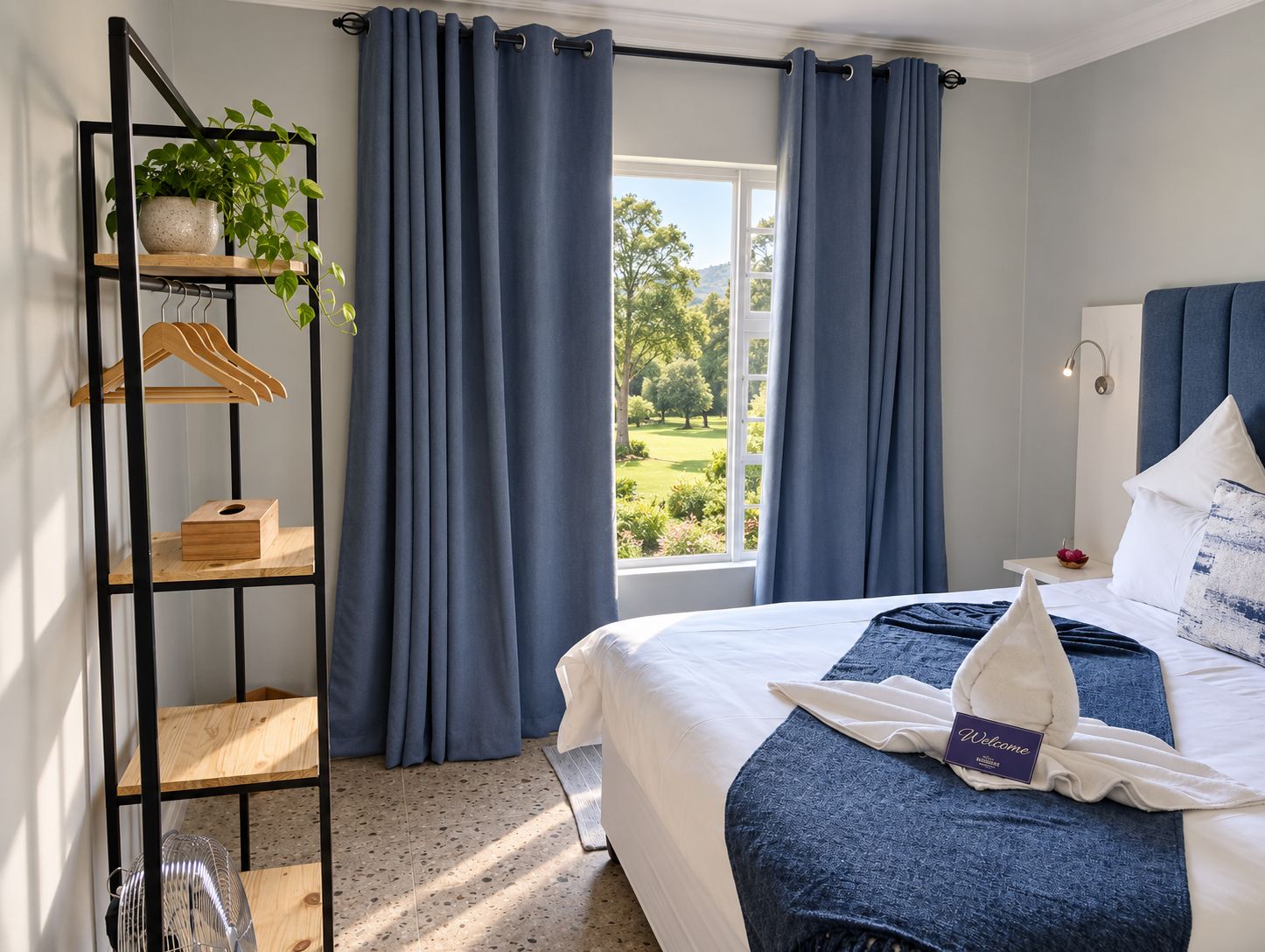 A hotel room with a bed, a bedside table, a nightstand, a window with curtains, a plant on a shelf, and a view of greenery outside.