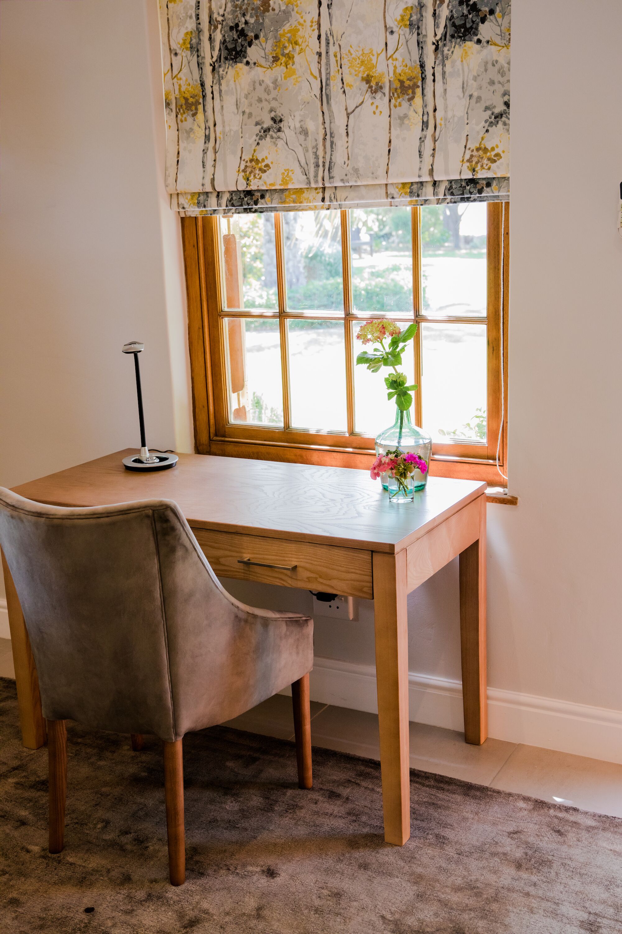 A wooden desk with a chair, a vase with flowers, and a lamp.