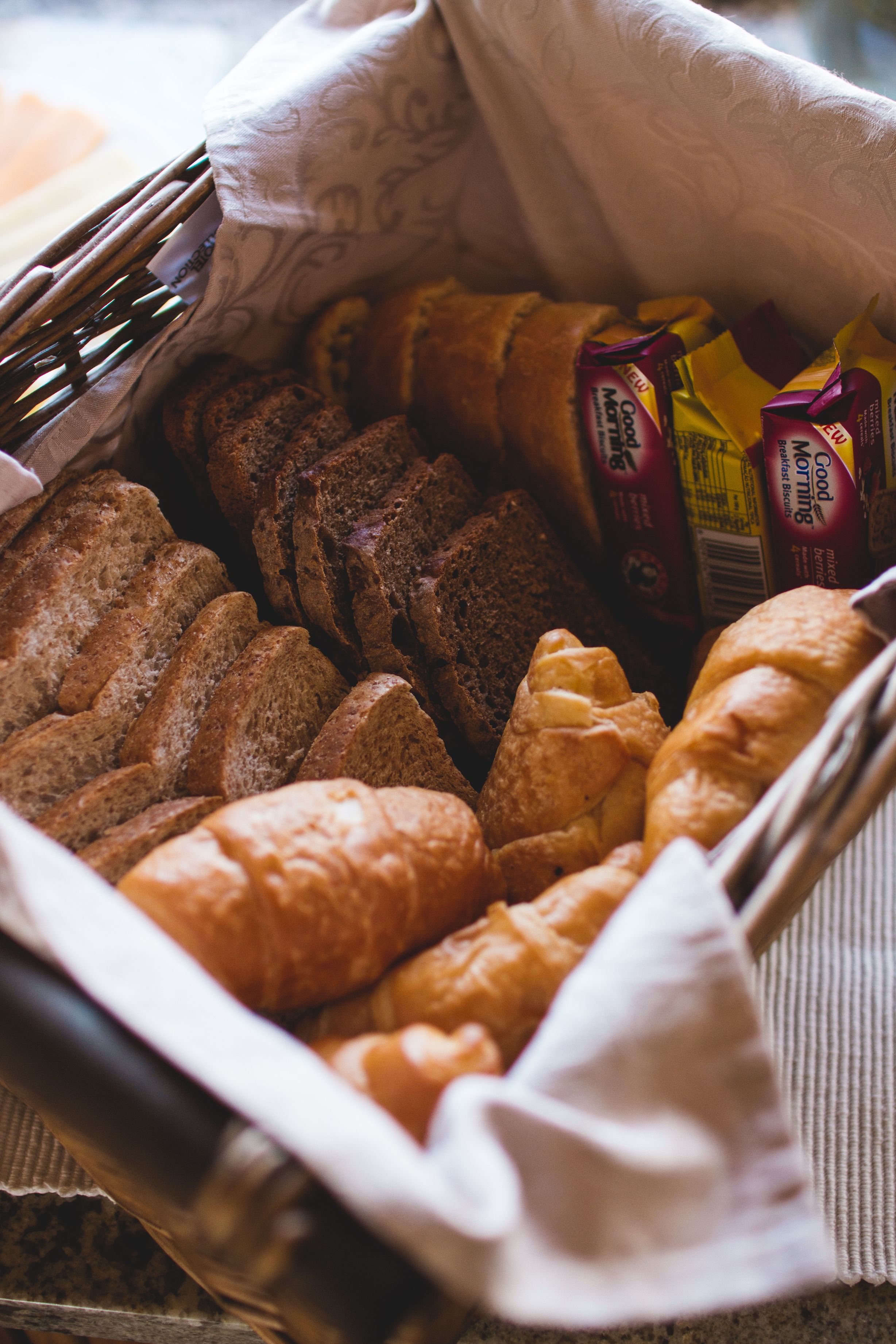 A basket filled with various types of bread and pastries, including sliced bread, croissants, and a bag of Good Morning brand coffee.