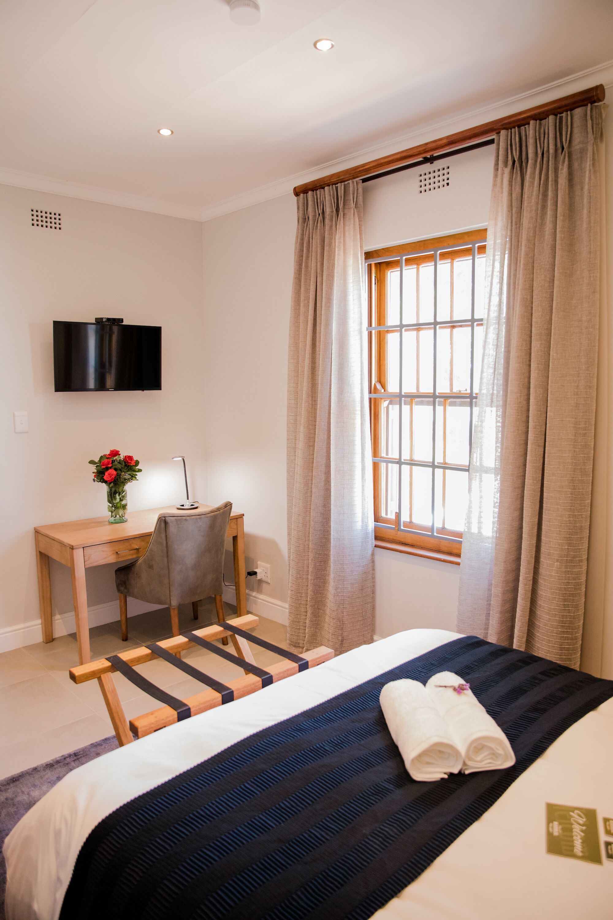 A hotel room with a bed, a desk with a chair, a TV mounted on the wall, and a window with curtains.