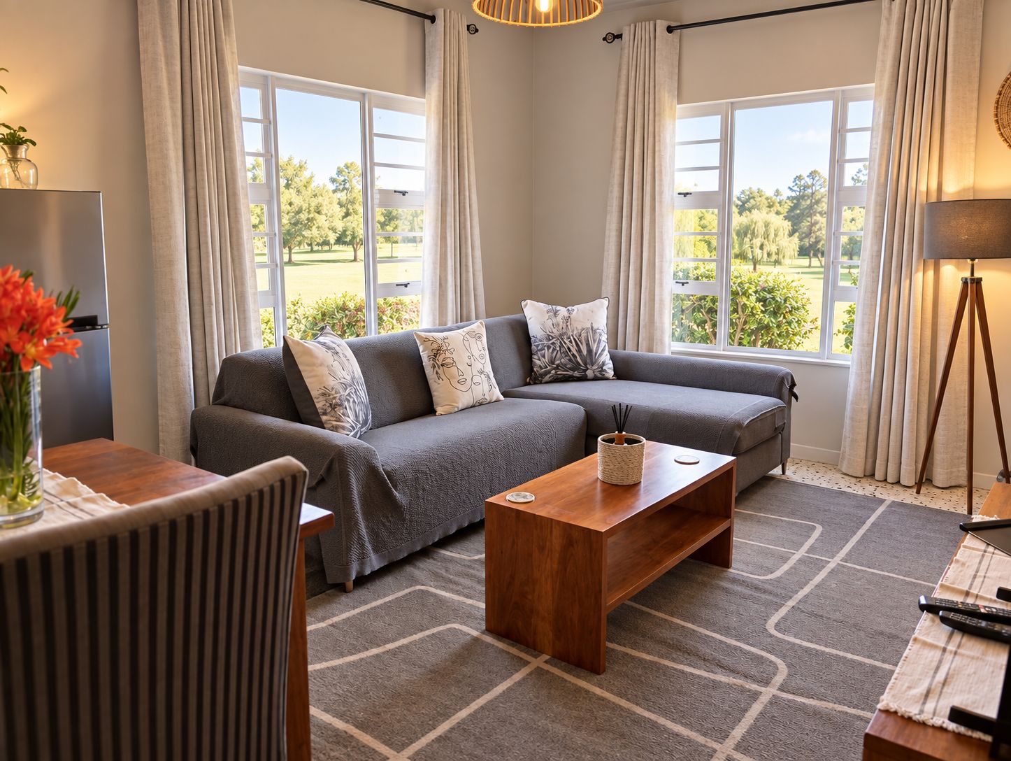 A modern living room with a grey sectional sofa, a wooden coffee table, and a view of a green landscape through large windows.