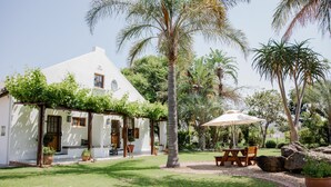 Una casa blanca con un porche cubierto, un patio con mesa y sillas, y vegetación tropical circundante.