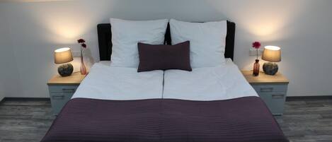 Una cama doble con cabecera oscura, dos mesitas de noche con lámparas y una colcha de color oscuro.