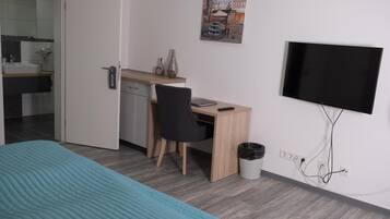 A hotel room with a bed, a desk, a chair, a TV, and a small trash bin.