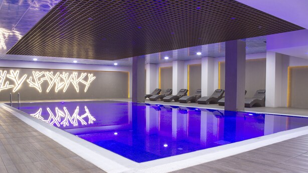 Piscina interior moderna con fuente iluminada en azul, tumbonas y diseño de techo geométrico.