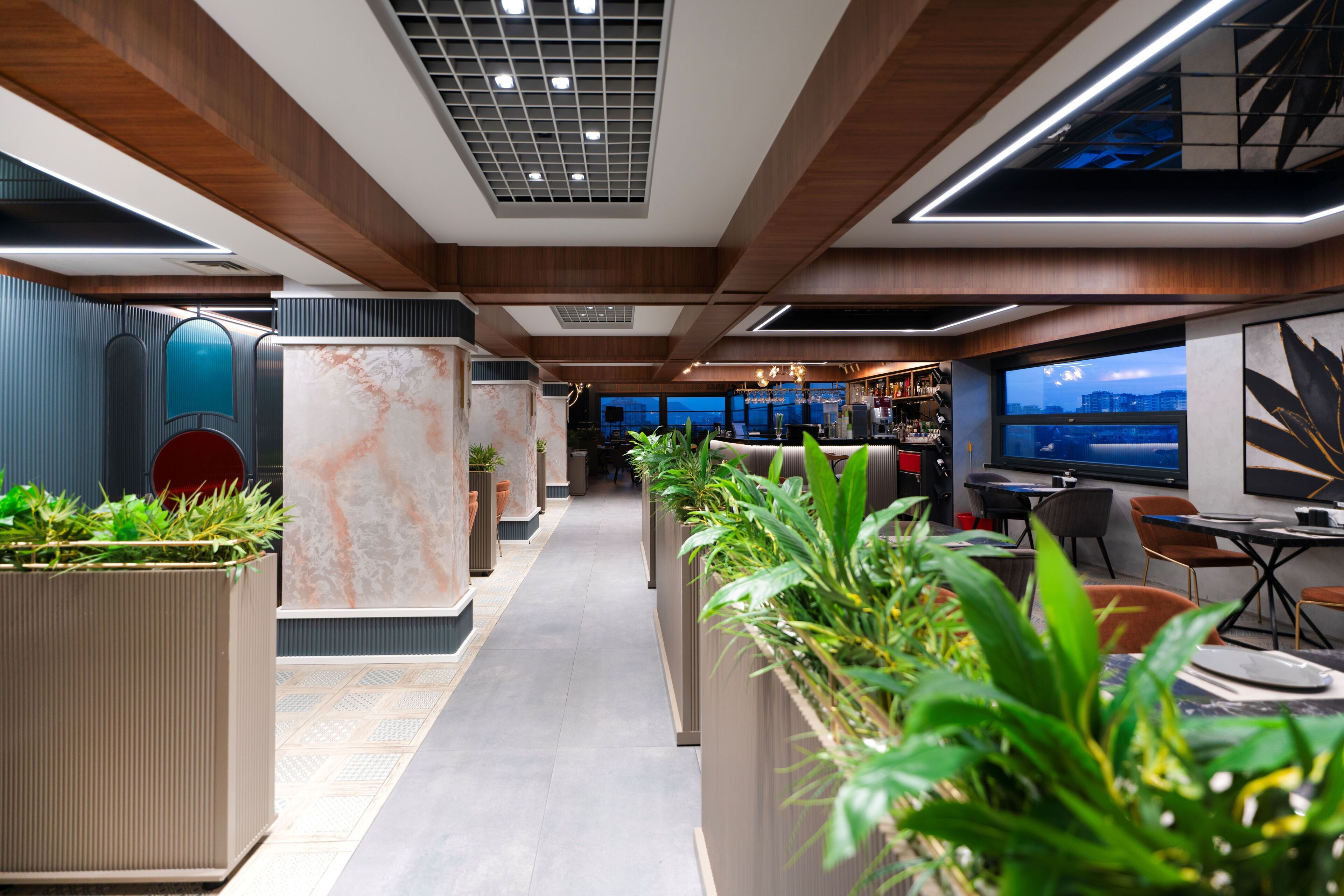 A modern interior with a long corridor lined with planters and seating areas, leading to a bar and a large window with a city view.
