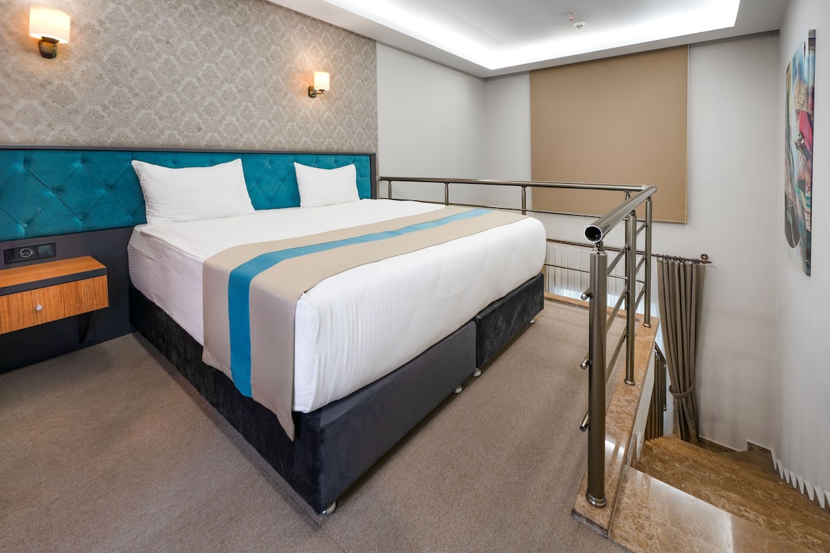 A modern hotel room with a large bed, a wooden nightstand, a headboard with a tufted design, a wall-mounted light fixture, and a view of the outside through a window.