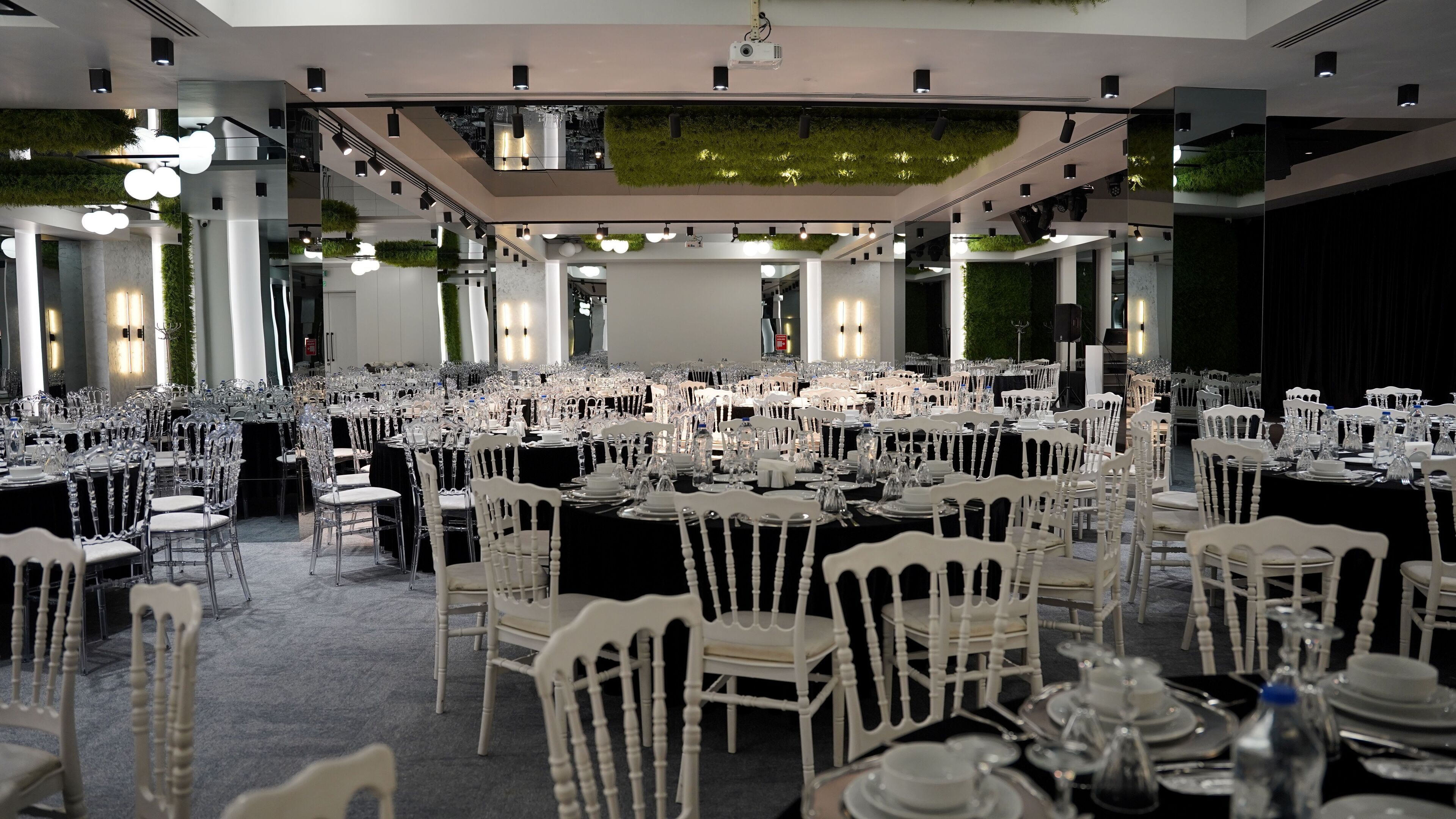 A large banquet hall with multiple round tables set for dining, white chairs, and a ceiling with recessed lighting and greenery.