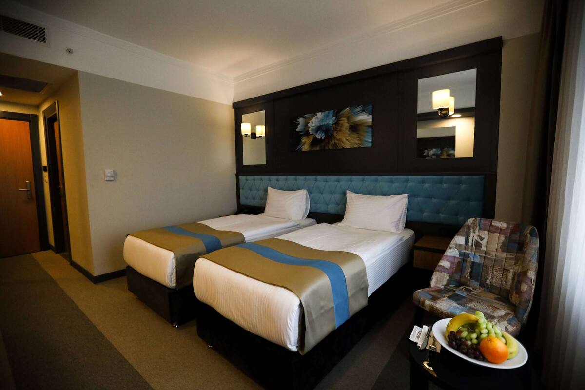 A hotel room with two beds, a blue upholstered headboard, a small table with fruit, and a chair with a patchwork design.