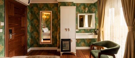 A hotel room with a wooden door, a bed, a green armchair, a small table, a mirror, a dresser, and a window with curtains.