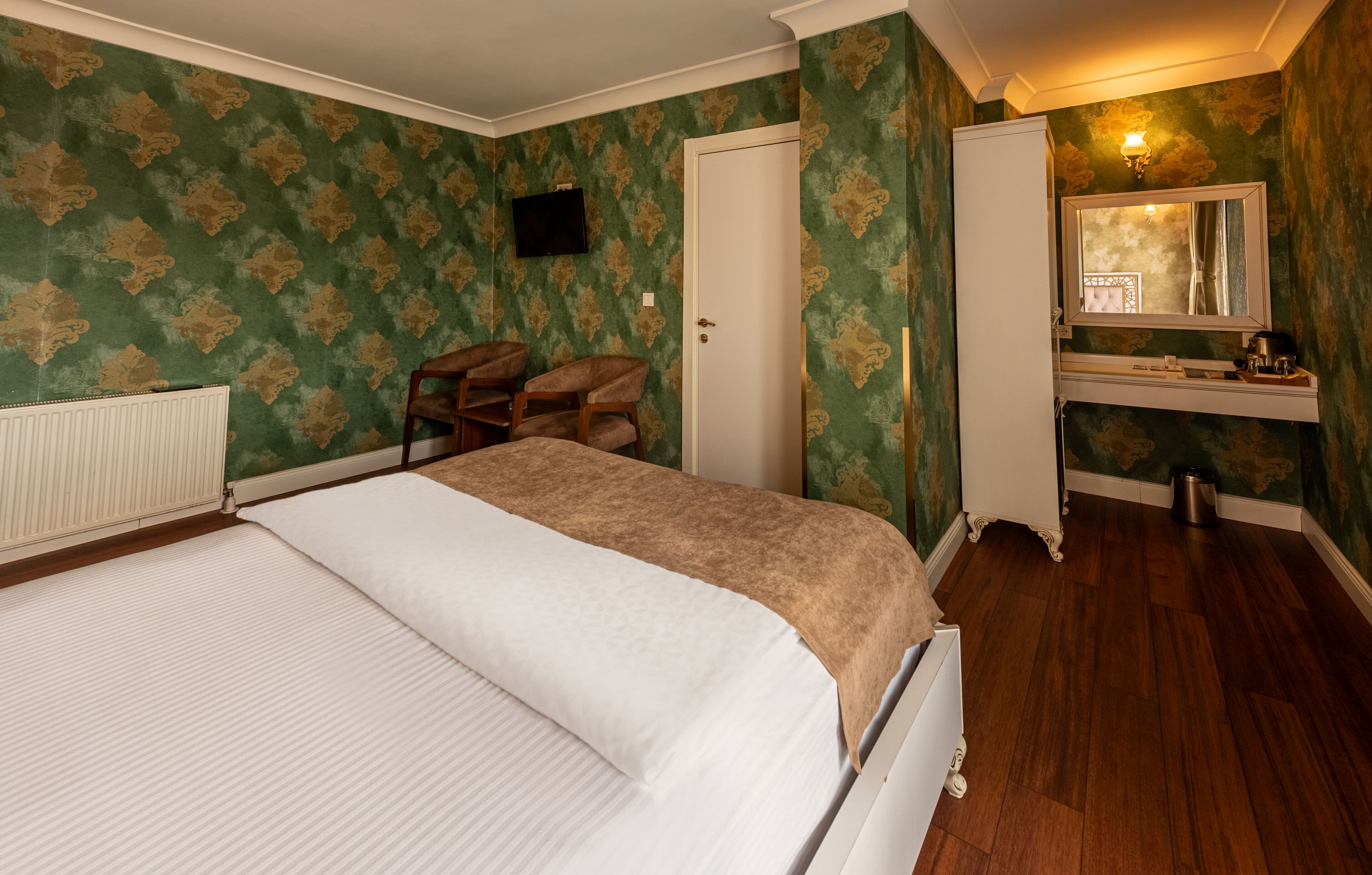 A bedroom with a bed, a chair, a vanity with a mirror, and a radiator.