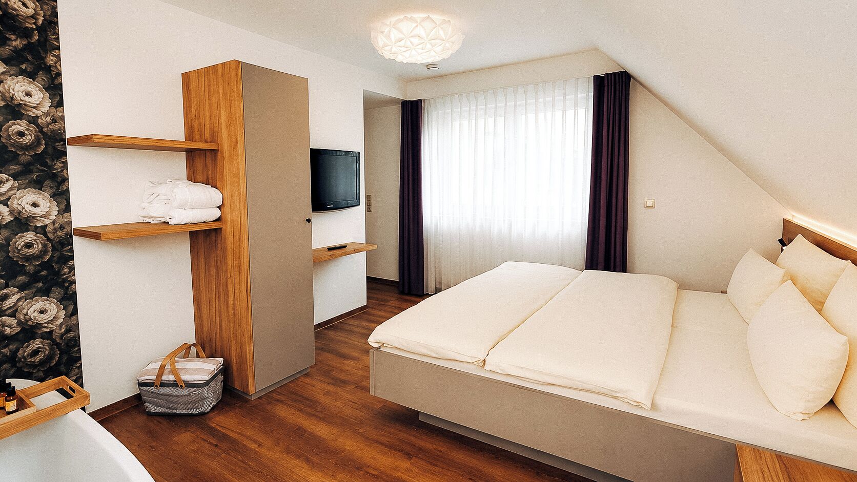 Habitación de hotel con cama, televisor, estante de madera, cesta y ventana con cortinas.