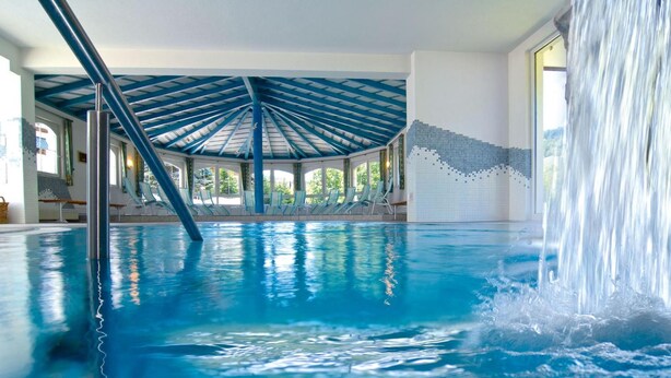An indoor swimming pool with a blue ceiling, a waterfall feature, and lounge chairs along the poolside. - Hotel Garni Bergblick (Biberach)