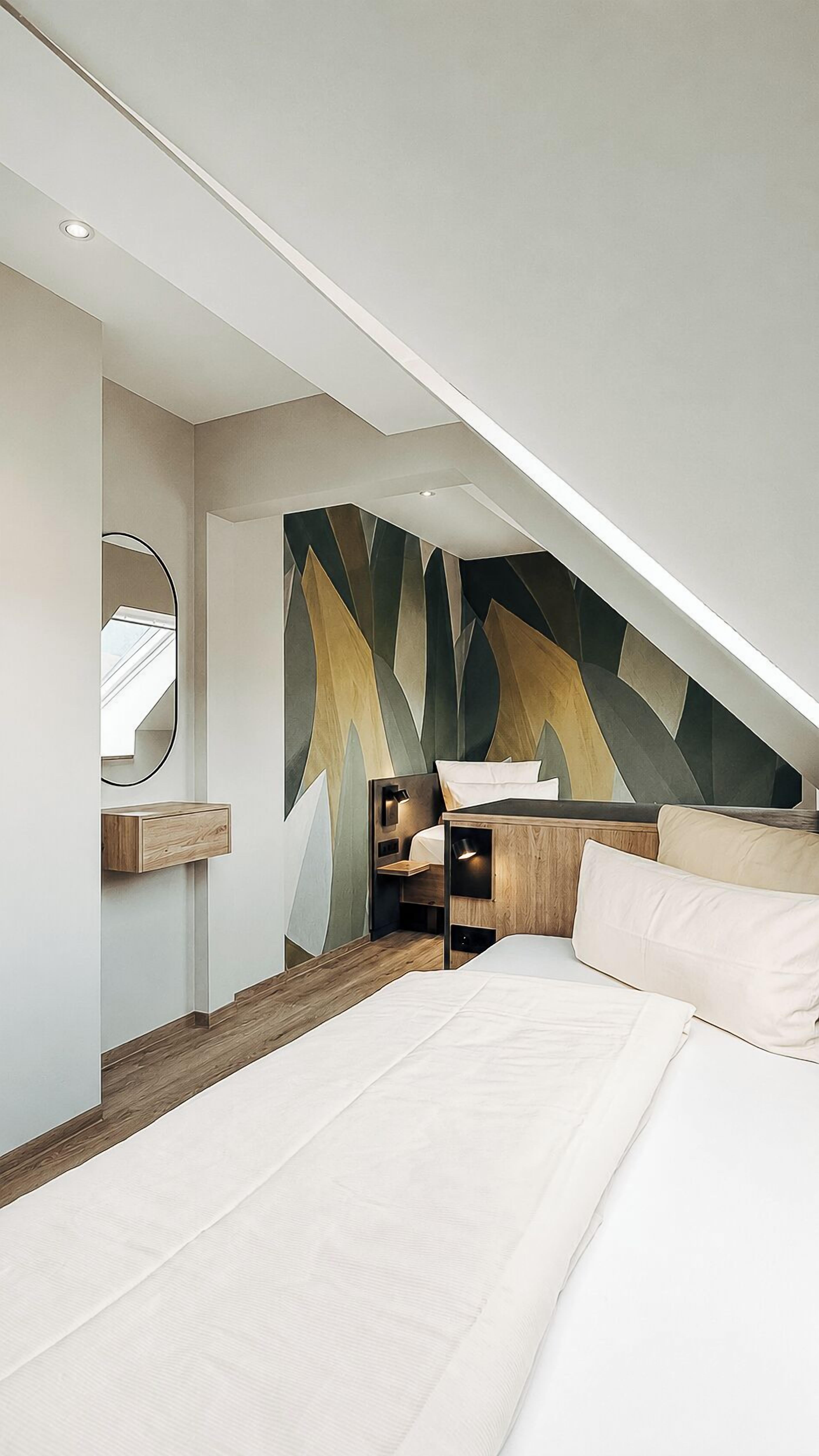 A modern bedroom with a sloped ceiling, a bed, a bedside table, a mirror, and a geometric-patterned wall.