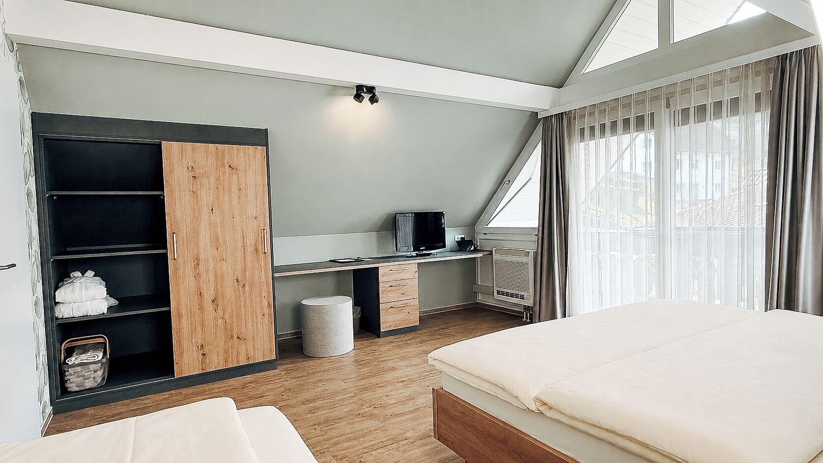 A bedroom with a bed, a desk with a computer, a wardrobe, and a window with curtains.