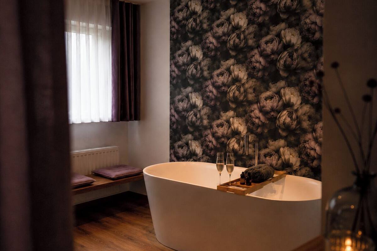 A modern bathroom with a freestanding bathtub, a window with curtains, a radiator, and a floral wallpaper.
