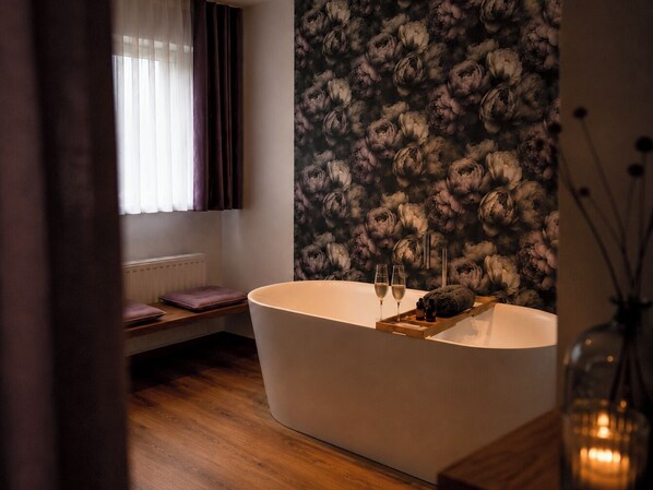 Un baño moderno con una bañera independiente, un ventanal con cortinas, un radiador y un papel pintado floral.