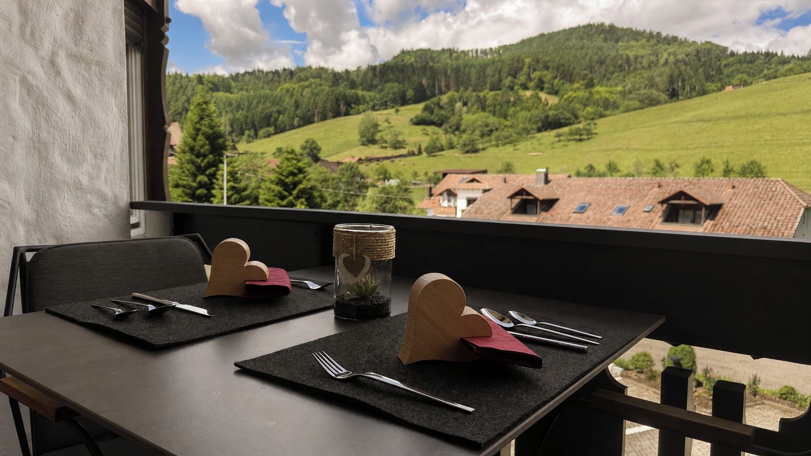 Une table à manger dressée avec des couverts et un porte-serviettes en forme de cœur, donnant sur un paysage verdoyant parsemé de maisons.
