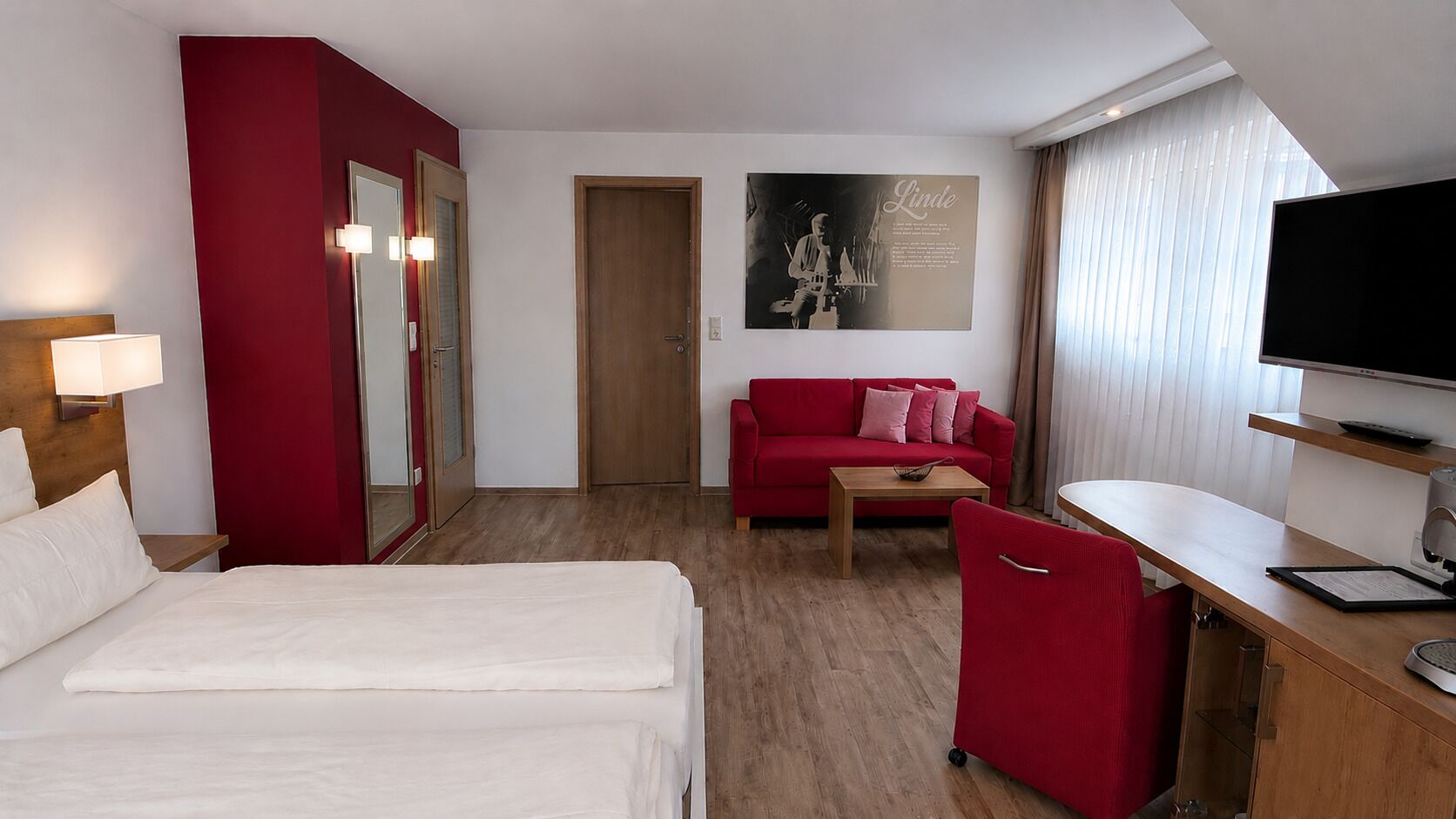 Une chambre d’hôtel avec un lit, un canapé rouge, un bureau, une télévision et un miroir.