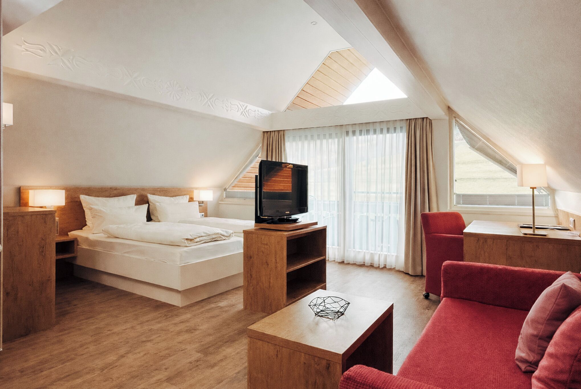 Habitació d'hotel moderna amb un llit, una televisió, un sofà vermell i una taula de centre de fusta.