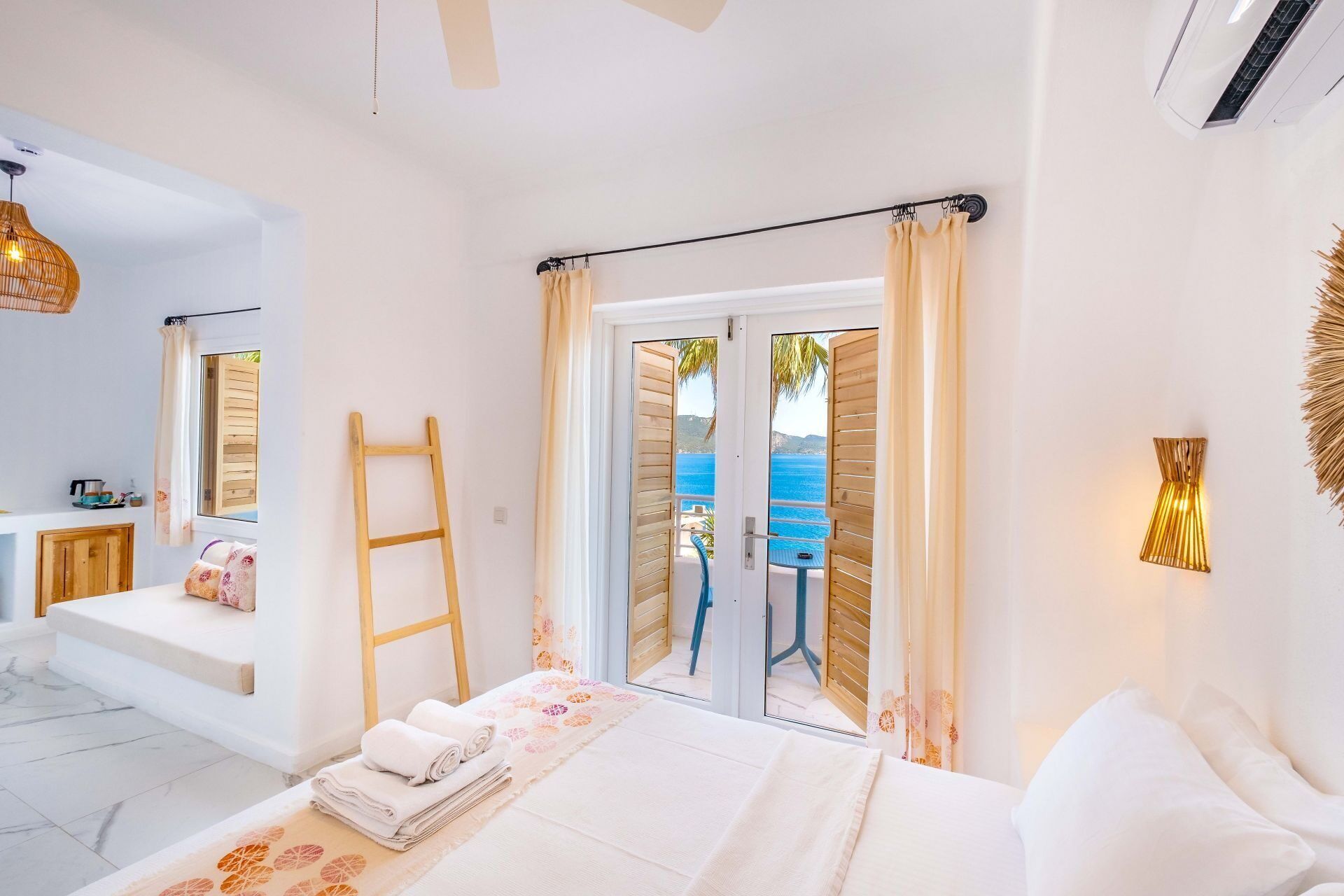 A bedroom with a bed, a wooden ladder, a view of the sea through a glass door, and a bathroom with a sink and mirror.