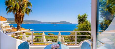 A balcony with a table and chairs, overlooking a coastal view with a palm tree and a mountain range in the distance.