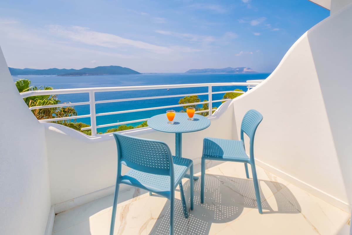 A balcony with two blue chairs and a small round table, overlooking a clear blue sea and distant mountains.