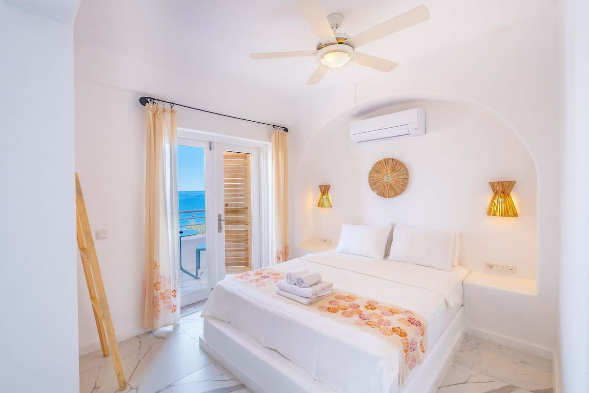 A bedroom with a bed, a ceiling fan, an air conditioning unit, and a view of the sea through a door.