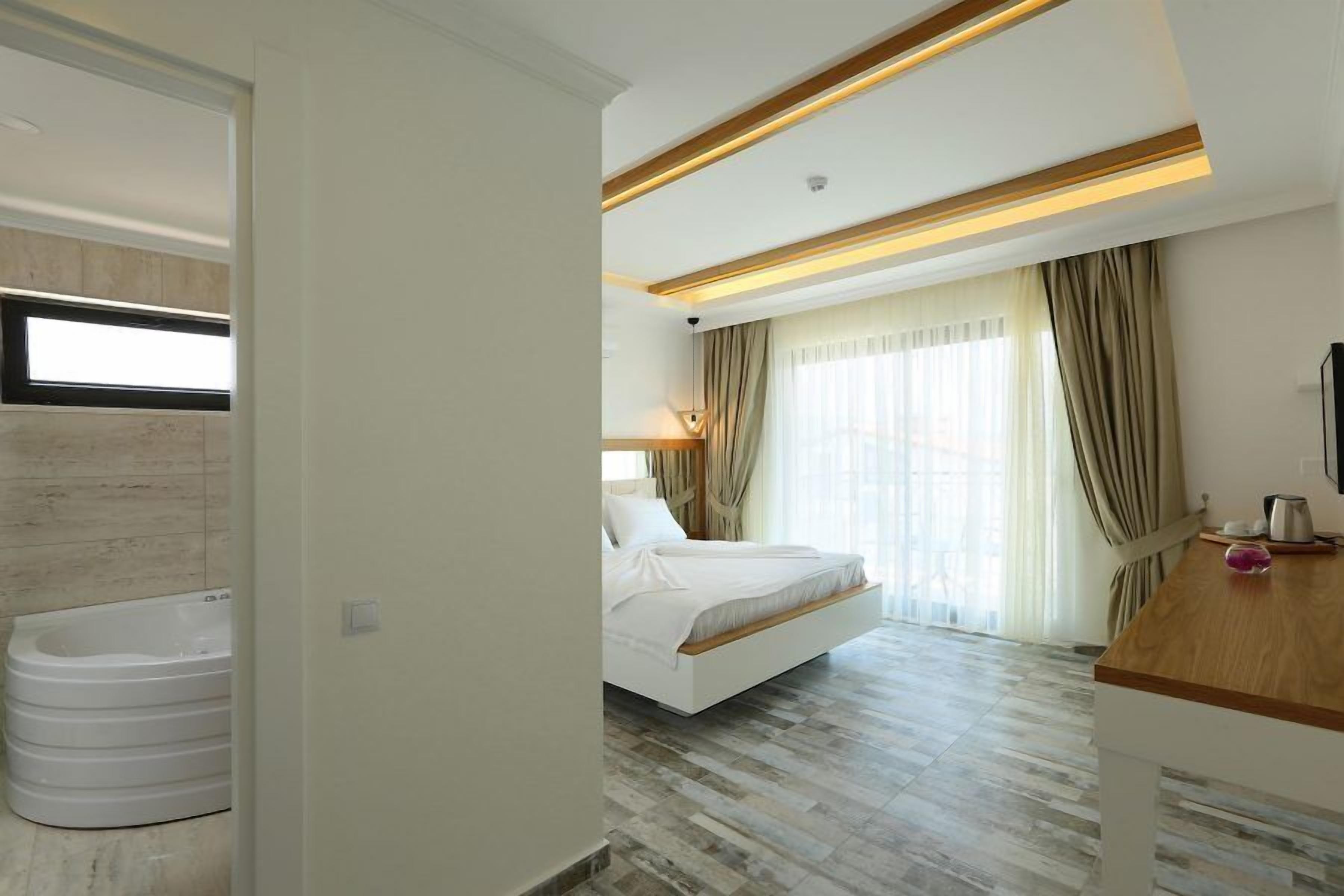Een moderne hotelkamer met een groot bed, een houten nachtkastje, een inbouwbad en een groot raam met gordijnen.