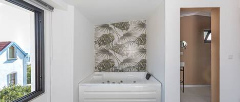 A modern bathroom with a large bathtub, a window with a view of a house, and a wall with a tropical leaf pattern.